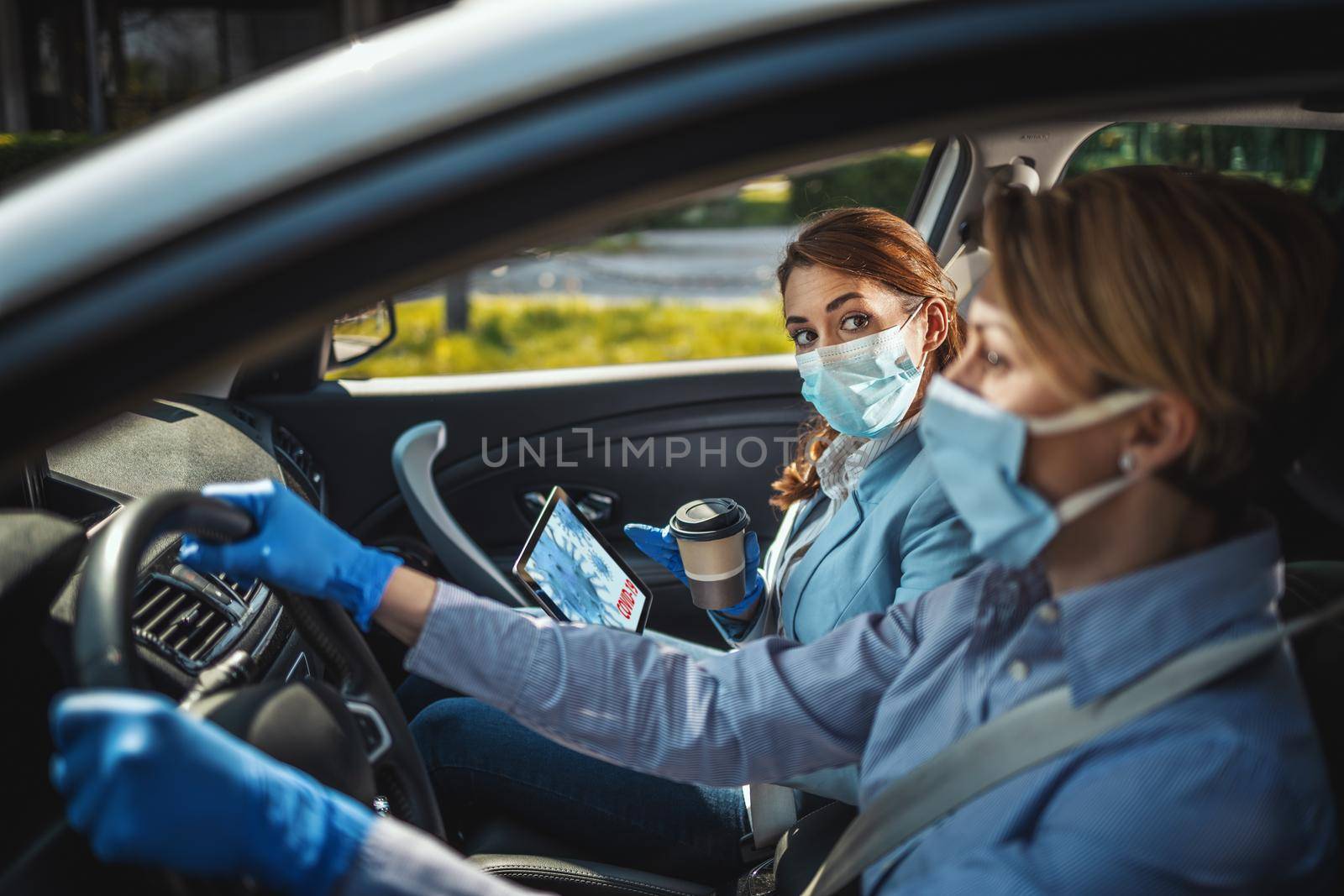 Young business women with protective masks on their faces are sitting in a car and talking about global crisis. They are showing each other an animation of a corona virus on a tablet.