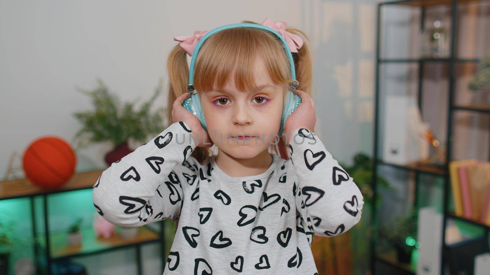 Close-up portrait of little happy child girl dancing on sofa while listening music in headphones in cozy living room. Small cute teen school kid at home alone dancing, singing and moving to the rythm