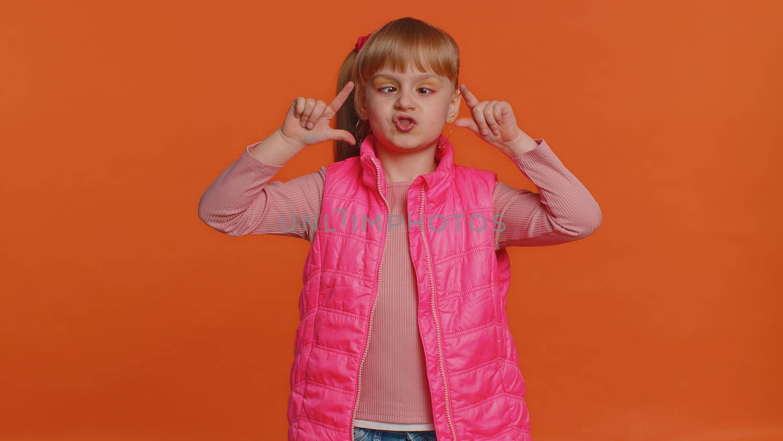 Funny joyful sincere little girl 6 years old in vest making playful silly facial expressions and grimacing, fooling around showing tongue. Young toddler child isolated alone on orange wall background