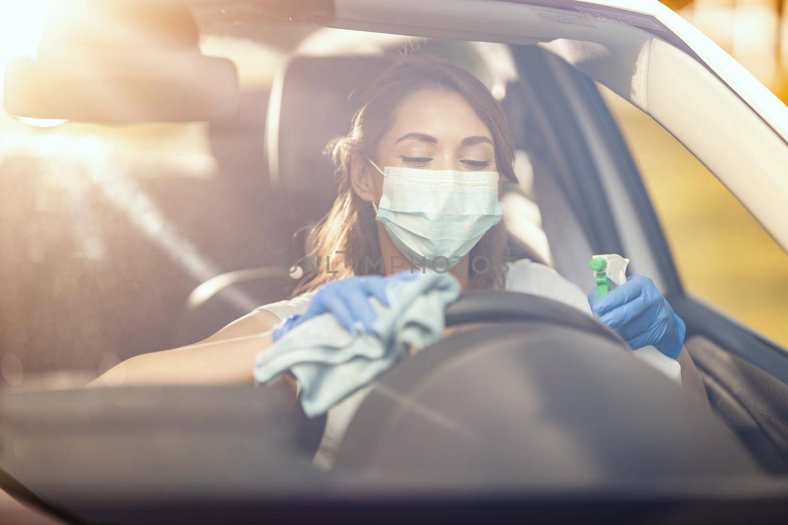 A young woman with a mask on her face  and protective gloves on her nands wipes her car holding a cloth in one and a bottle with disinfectant in the other hand.