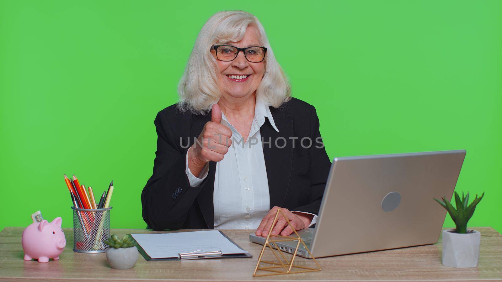 Senior old business woman employee using laptop computer notebook, raises thumbs up agrees with something or gives positive reply recommends advertisement likes good sits at green chroma key office