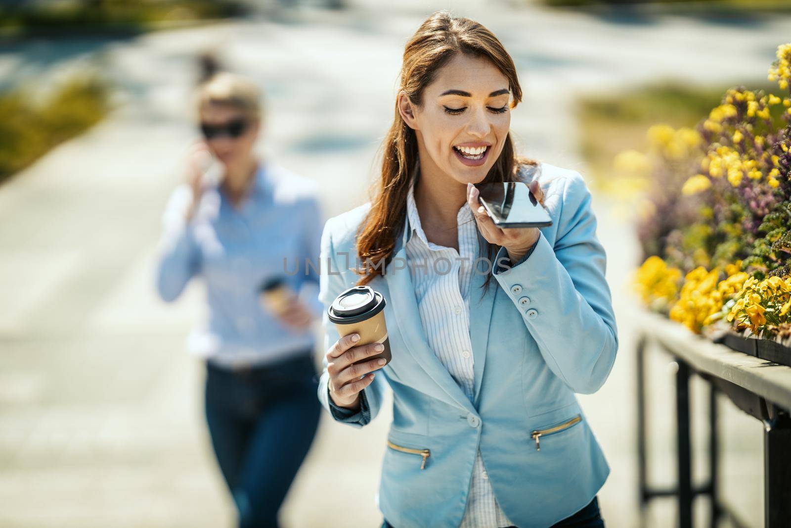 Young smiling businesswoman is walking in front of office and sending voice message during coffee break.