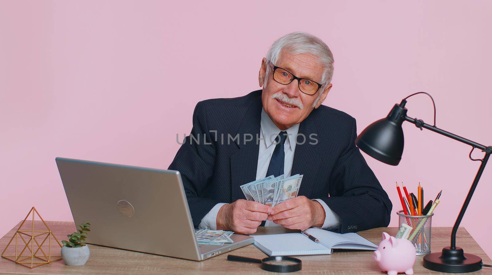 Senior business man accountant or banker making calculations at workplace desk. Elderly grandfather counting money dollar cash banknotes income earnings, working at office on pink studio background