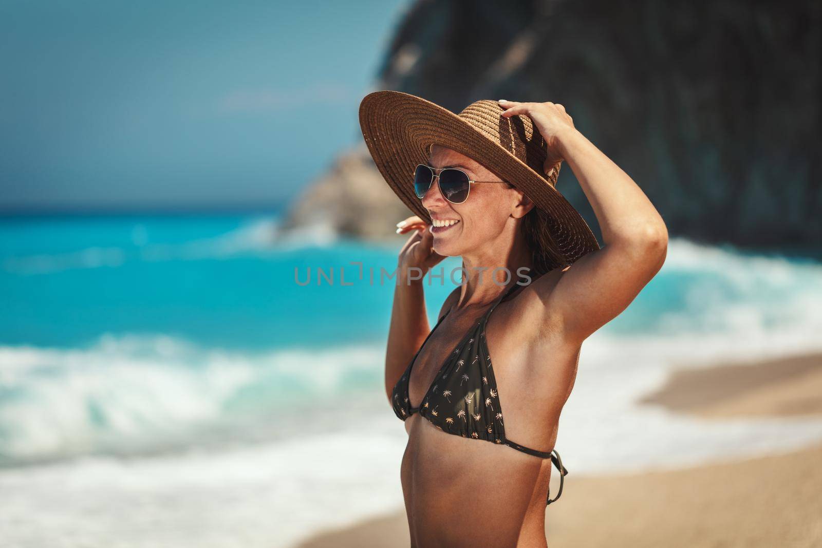 Handsome midle age woman in bikini wearing straw hat is walking along the beach and enjoying view on ocean at summer sun.