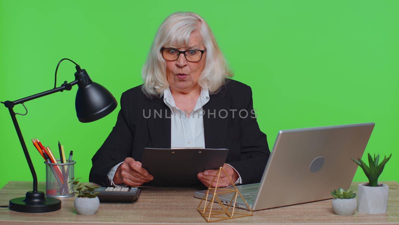 Oh my God Wow. Senior business woman, using laptop, analysing paperwork documents raising hands in surprise, shocked by sudden victory, good news. Elderly freelancer working at green chroma key office
