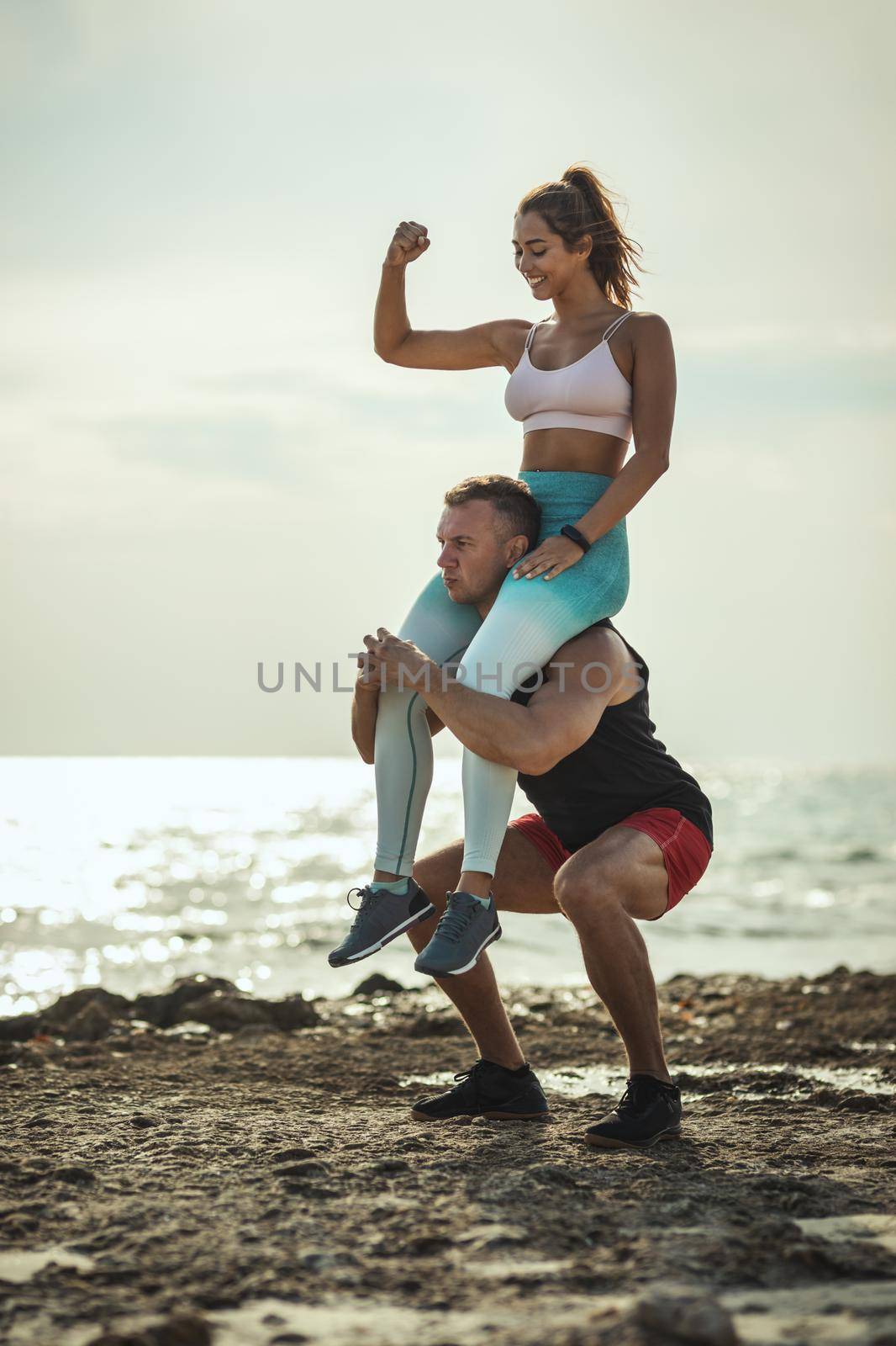 A handsome man is doing exercises on a sea beach with a girl on his shoulders.