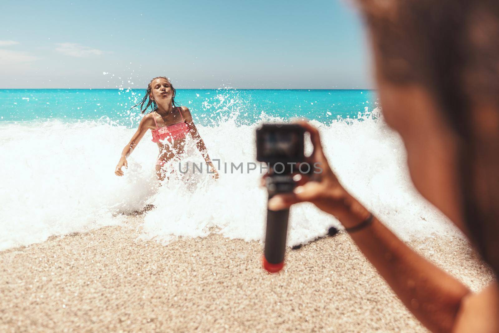 Woman with waterproof action camera takes a photo of her loving daughter in sea waves on the tropical beach. Selective focus. 