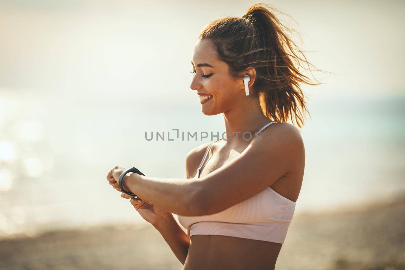 A beautiful young woman is looking at smartwatch and preparing to do jogging at the sea beach in summer sunny day.