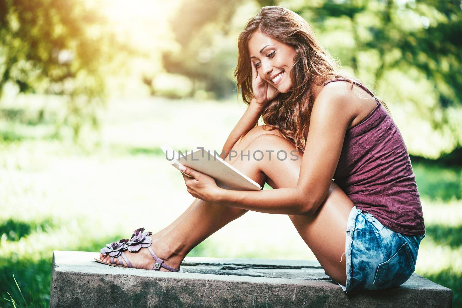 Attractive young woman is enjoying her time outside reading book in park with sunset in background.