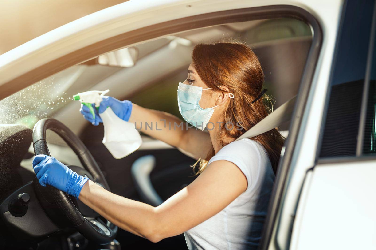 A young woman with a mask on her face  and protective gloves on her nands wipes a steering wheel in her car holding a bottle with disinfectant in hand.
