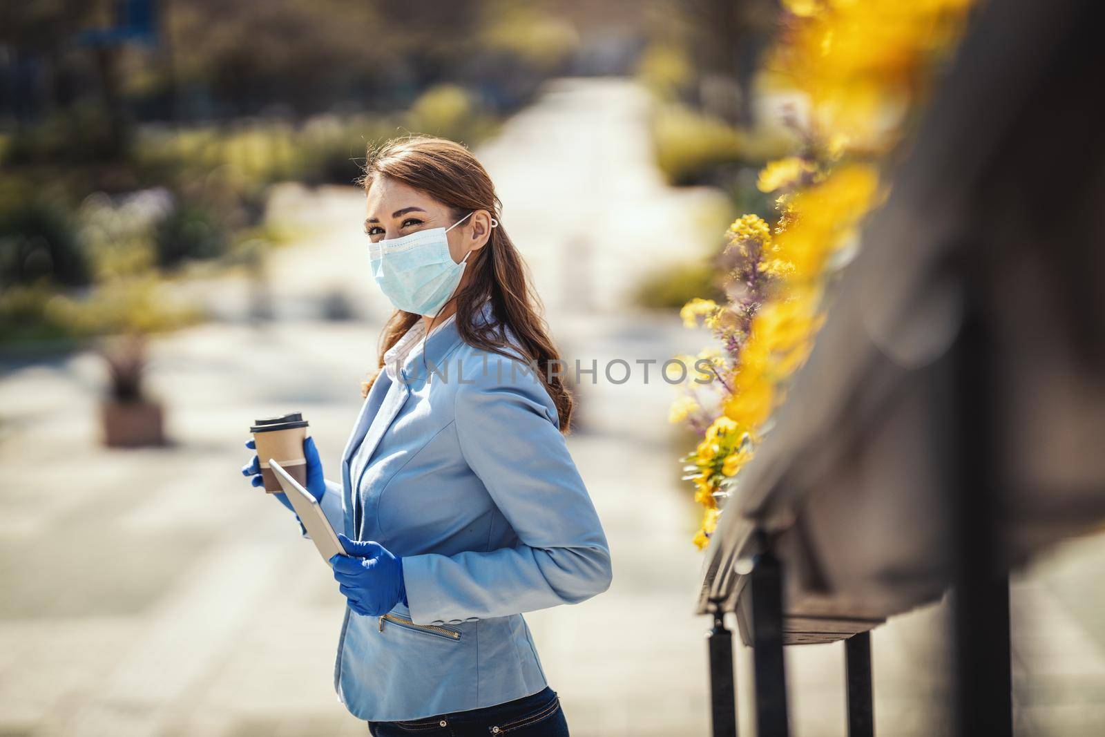 Young business woman with protective mask on her face and digital tablet in her hand is having coffee break outdoors.