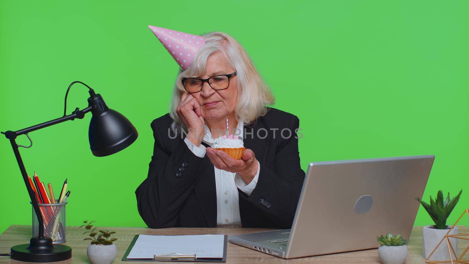 Sad upset lonely senior businesswoman in festive hat celebrating birthday party alone, feel frustrated despair disappointed sadness holds cake with candle at office. Mature woman on green chroma key