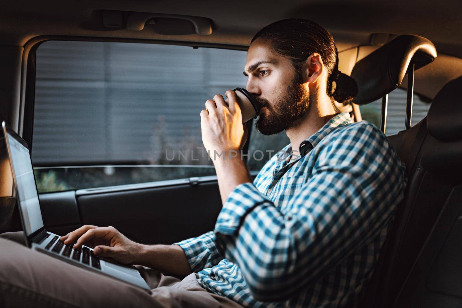 Young confident businessman is holding a cup of coffee in his hand, drinking and texting on a laptop on the back seat in car.