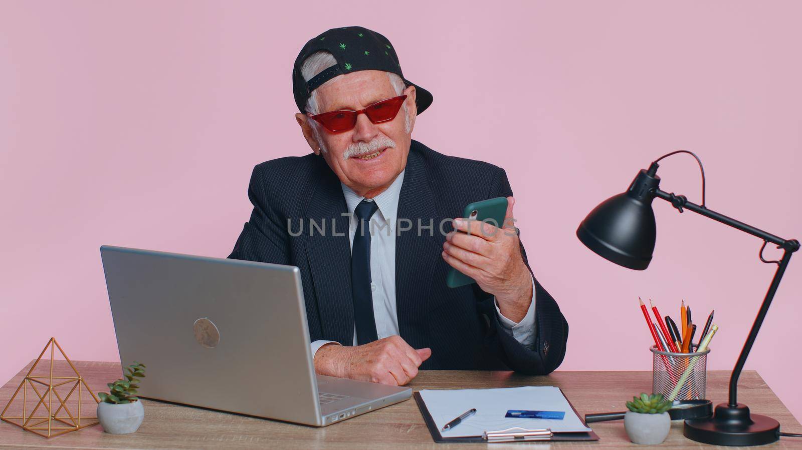 Funny stylish businessman using smartphone typing new post on web sms message, browsing, searching, addiction of social networks at office. Senior grandfather man studio isolated on pink background