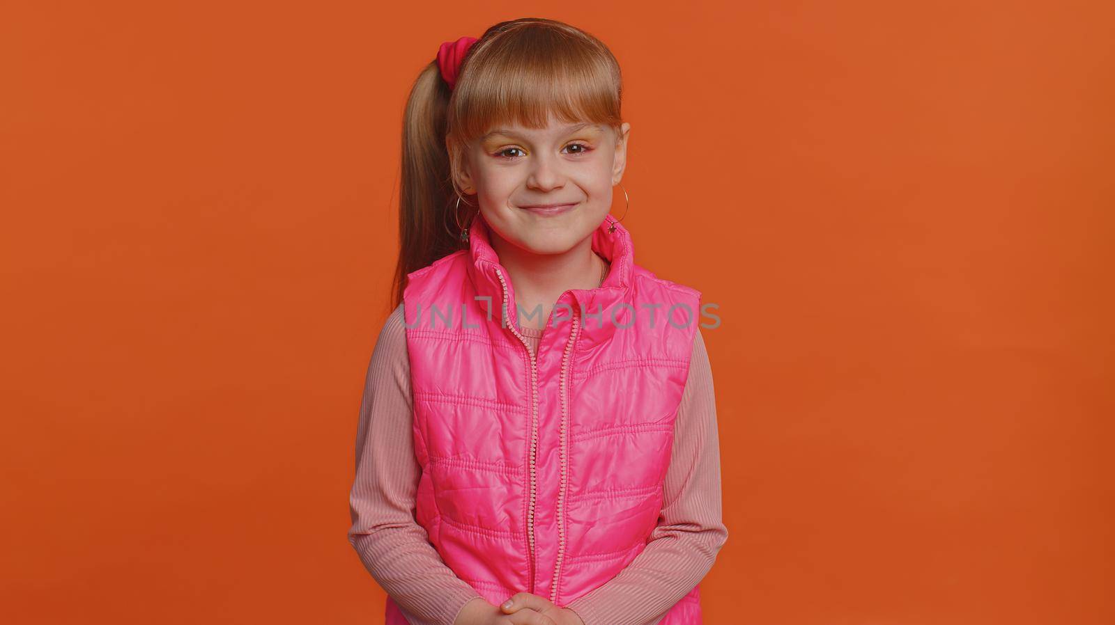 Cheerful happy toddler little girl wearing blouse, vest looking smiling at camera. Young fashion model child kid with brown eyes indoor studio shot isolated on orange background. Female nature beauty