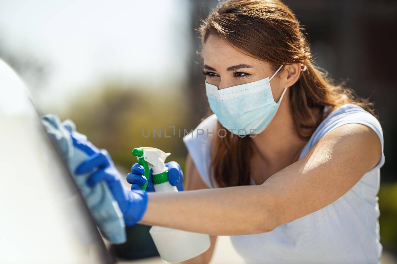 A young woman with a mask on her face and protective gloves on her hands, wipes her car holding a cloth in one and a bottle with disinfectant in the other hand.