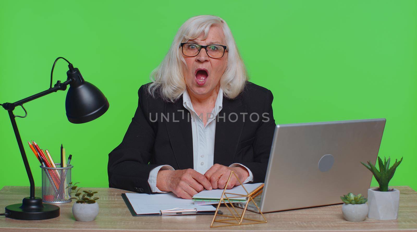 Upset senior businesswoman making face palm gesture, feeling bored, disappointed in result, bad news sits at workplace desk at office. Elderly mature woman isolated indoor on green chroma key studio