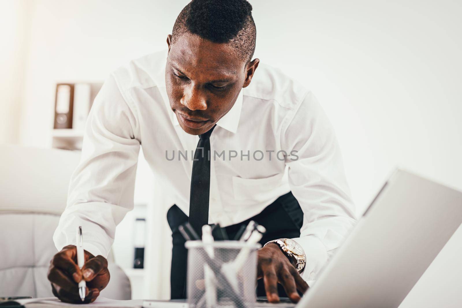 Pensive African businessman is writing notes in notebook and working on laptop in modern office.