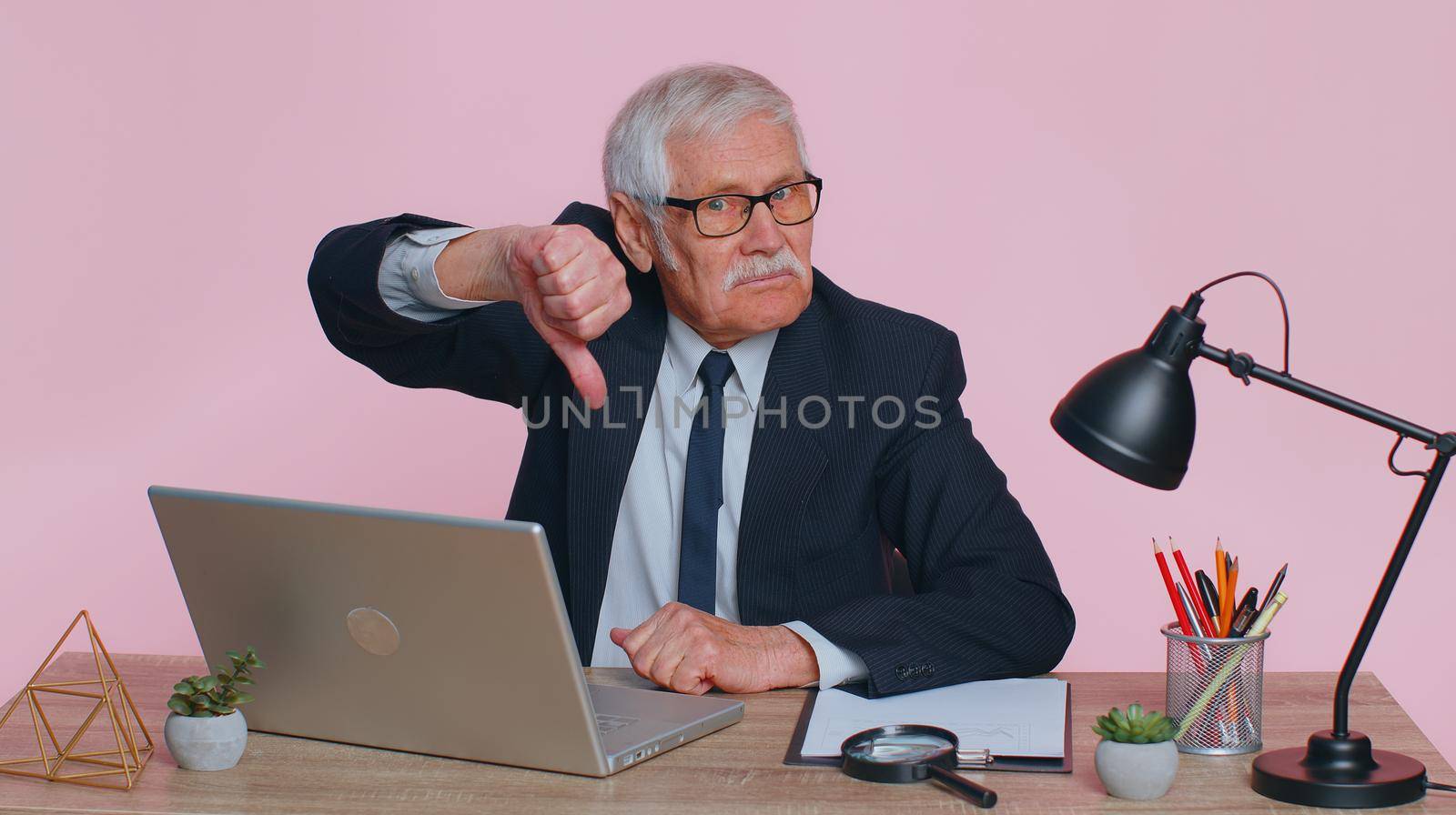 Upset senior mature business man showing thumbs down sign gesture, expressing discontent, disapproval, dissatisfied, dislike at pink office, using laptop computer. Elderly old grandfather freelancer