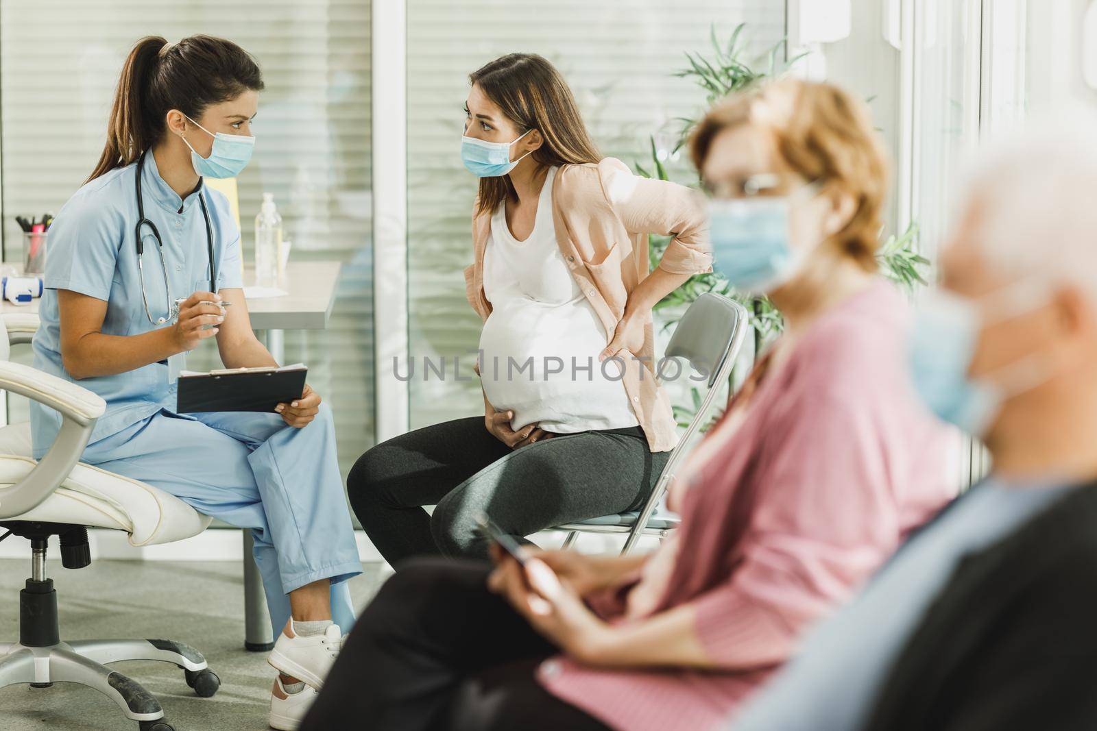 Pregnant Woman With Protective Mask Talking To Nurse At Waiting Room by MilanMarkovic78
