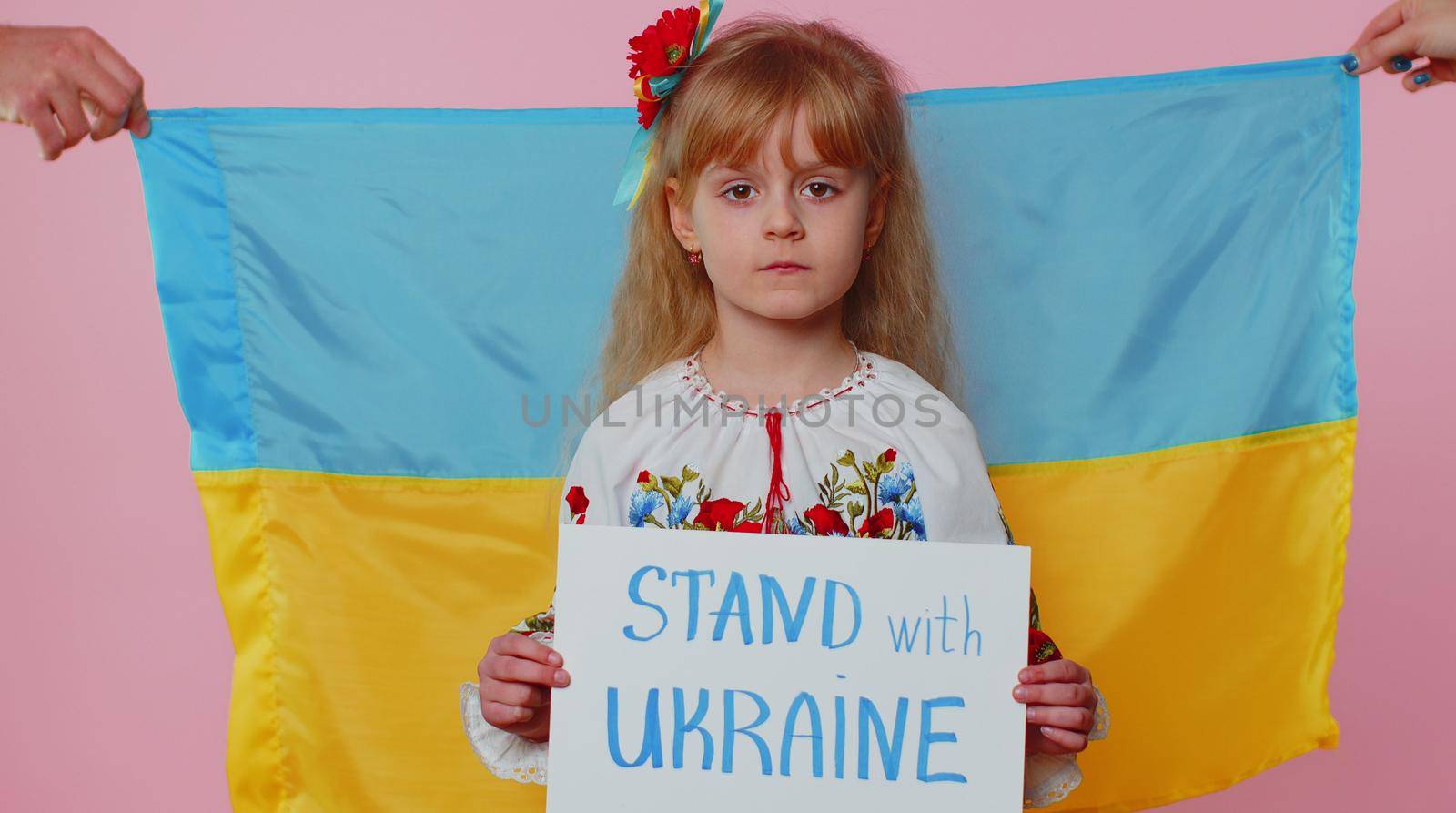 Upset toddler Ukrainian girl kid in embroidery dress protesting war conflict on pink background raises banner with inscription massage Be For The Truth Be With Ukraine. Peace, no war, stop aggression