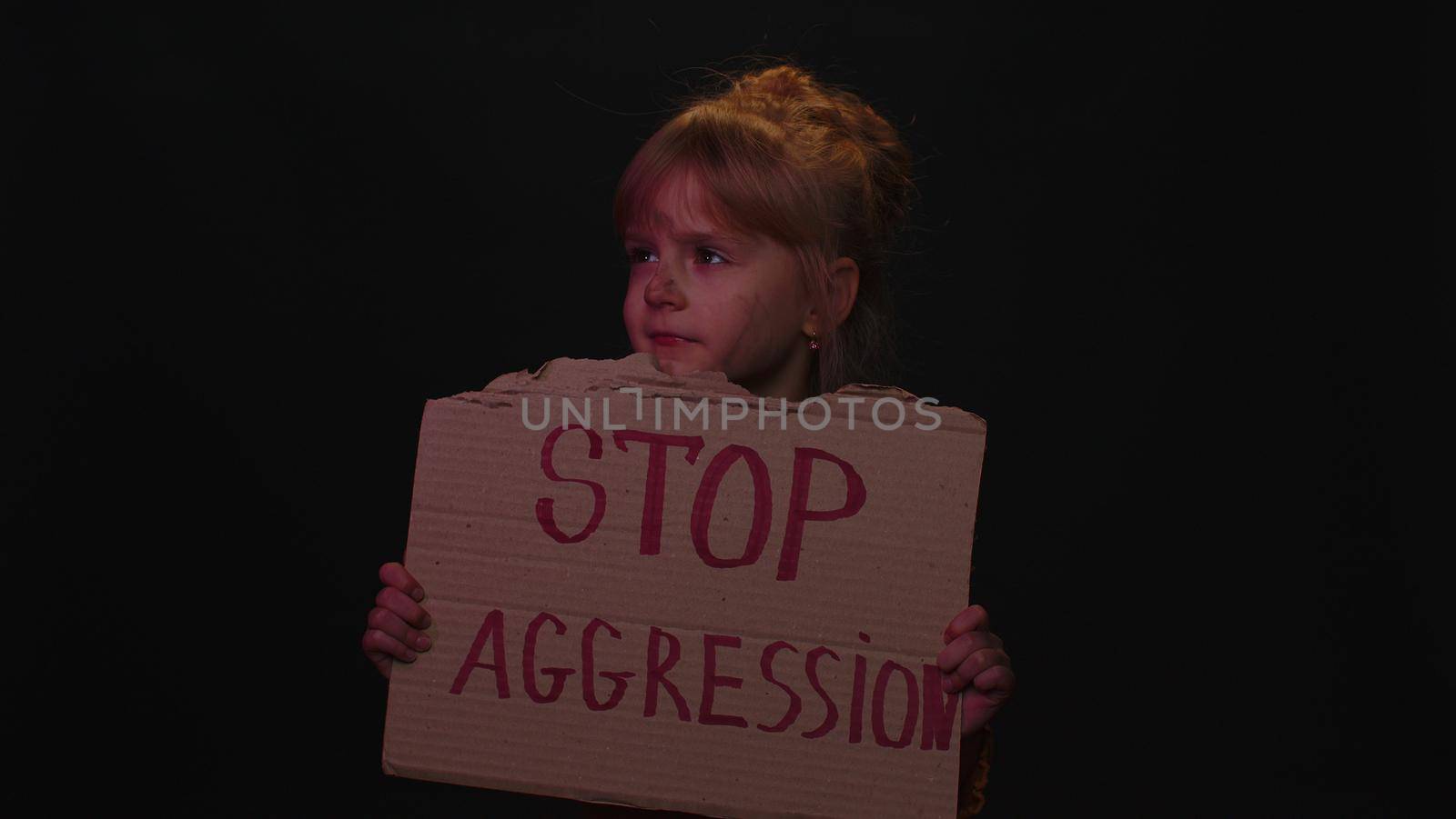 Upset afraid poor toddler child girl kid homeless with dirty face protesting war raises banner with inscription massage text Stop Aggression on black background. Crisis, peace, no war. Family conflict