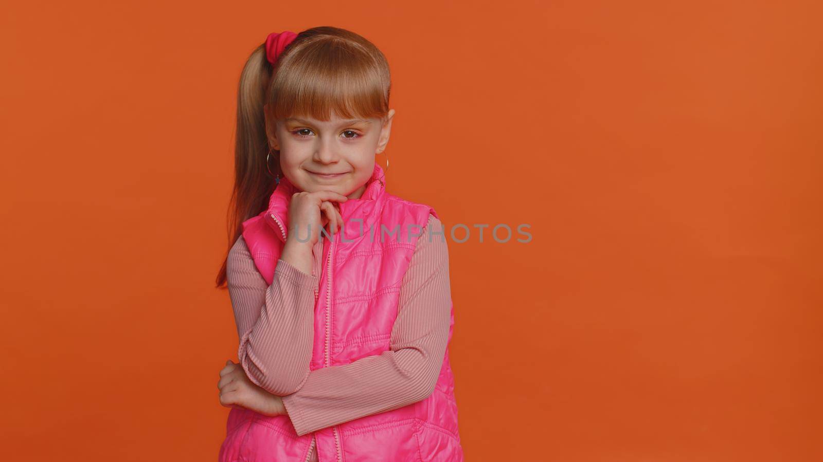 Cheerful lovely toddler little girl wearing blouse, vest looking smiling at camera. Young fashion model child kid with brown eyes indoor studio shot isolated on orange background. Female nature beauty
