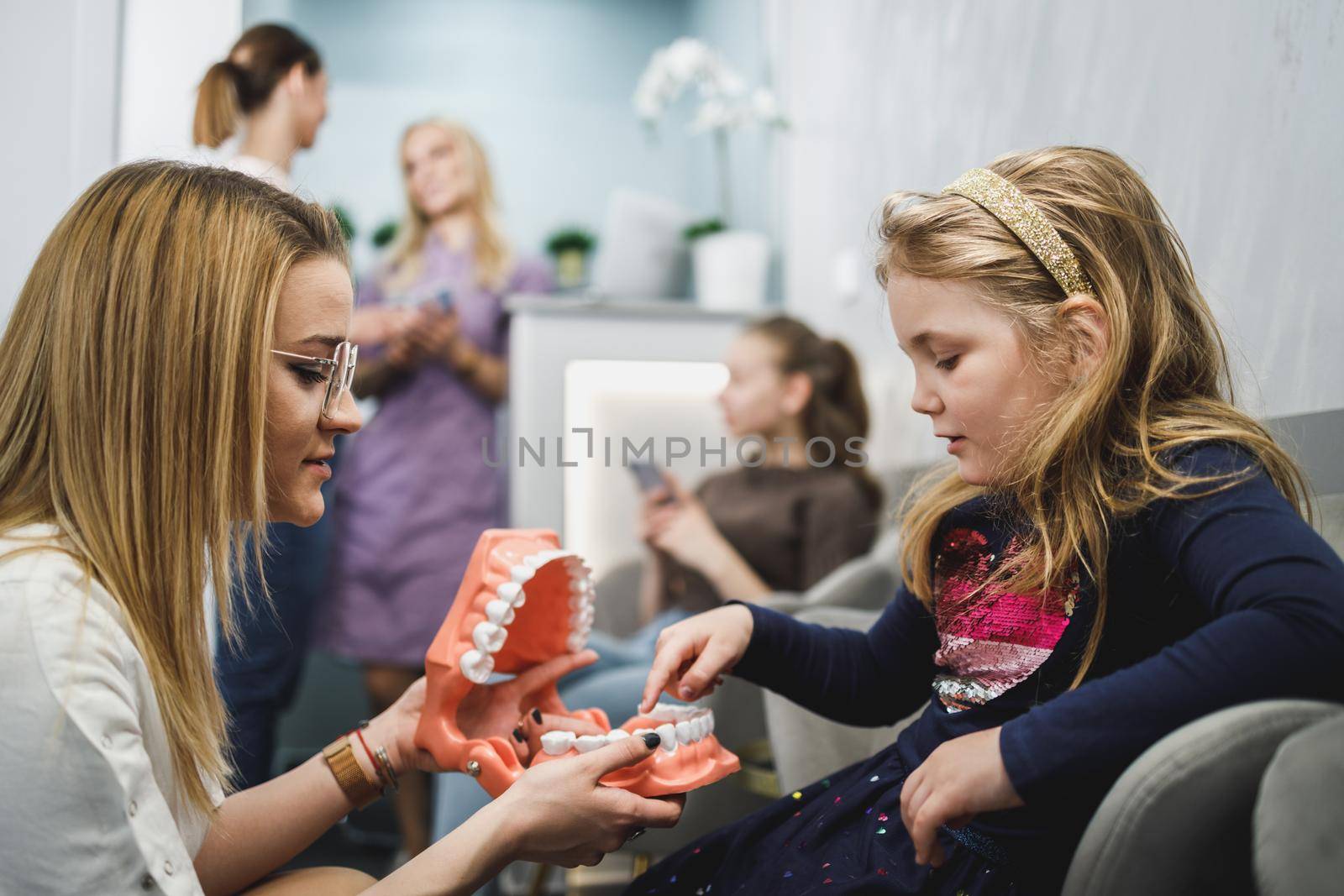 Female dentist and little girl holding model of teeth and communicating in waiting room at dentist's office.