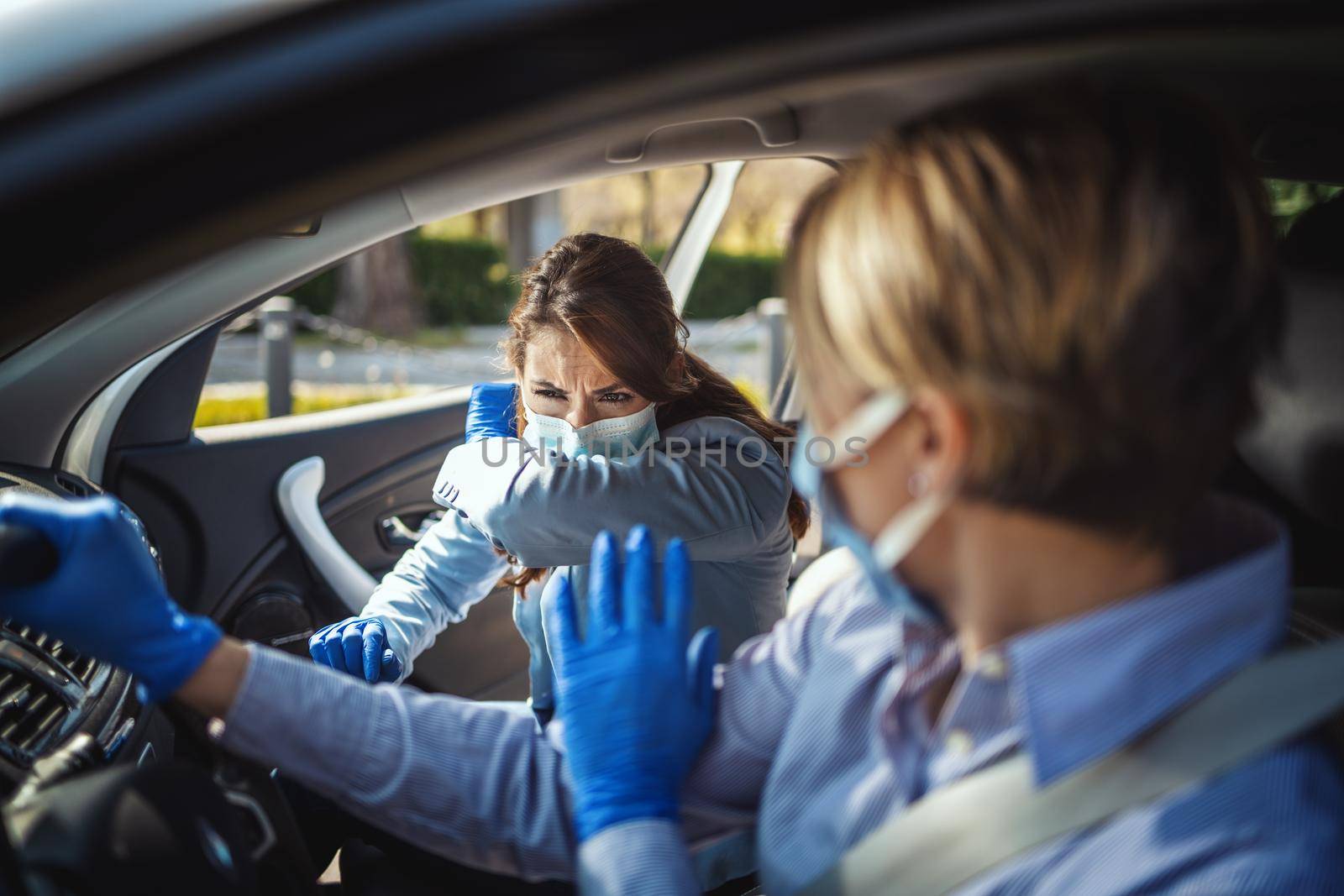 Young business women with protecting masks on their faces are sitting in a car. One of them is sneezing into the elbow try to avoid the spread of coronavirus.