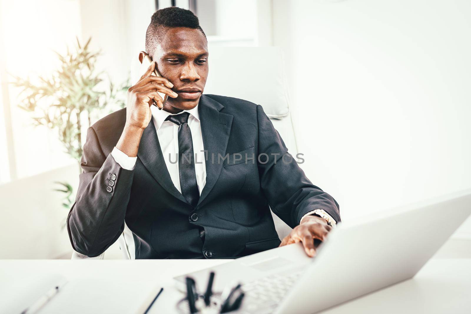 Pensive African businessman talking on smartphone and working on laptop in modern office.