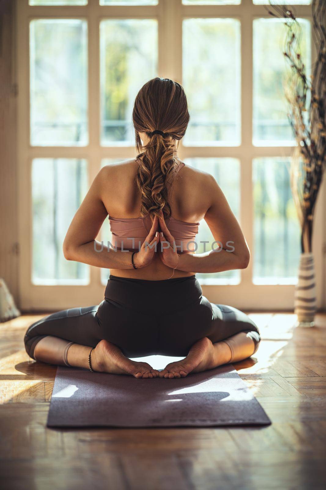 Young woman is doing yoga back namaste pose during coronavirus pandemic in the living room at home. She is meditating on floor mat in morning sunshine.