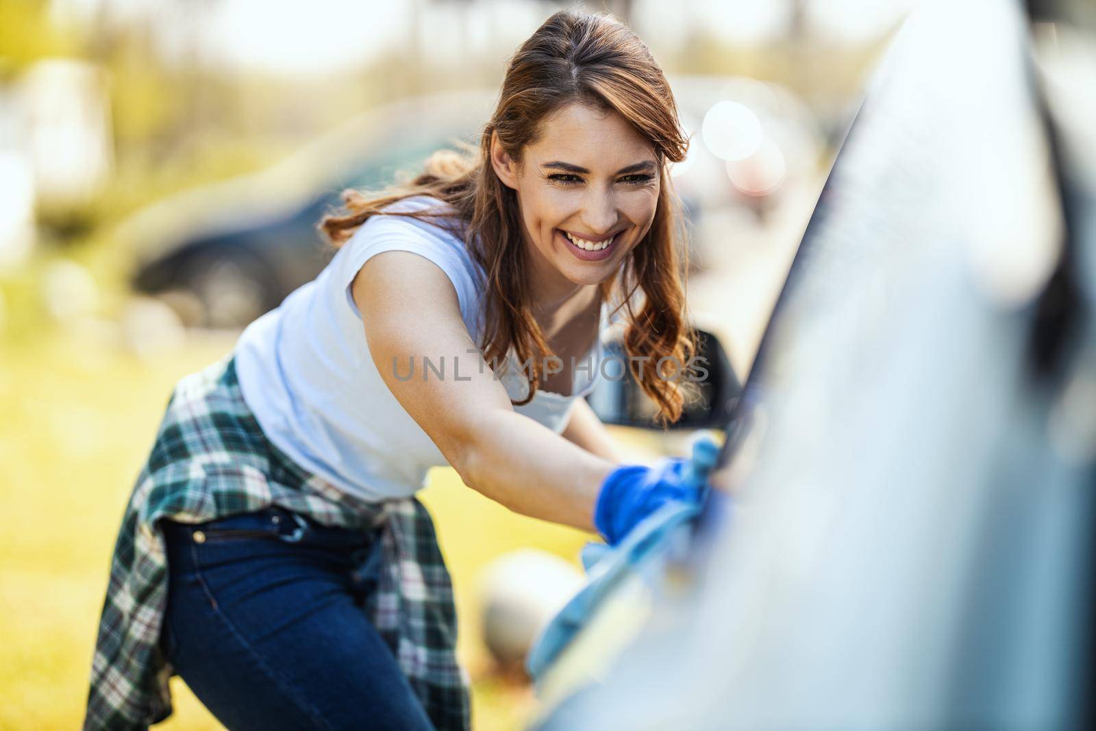 A smiling young woman with protective gloves on her nands wipes her car with a cloth ready to hit the road.