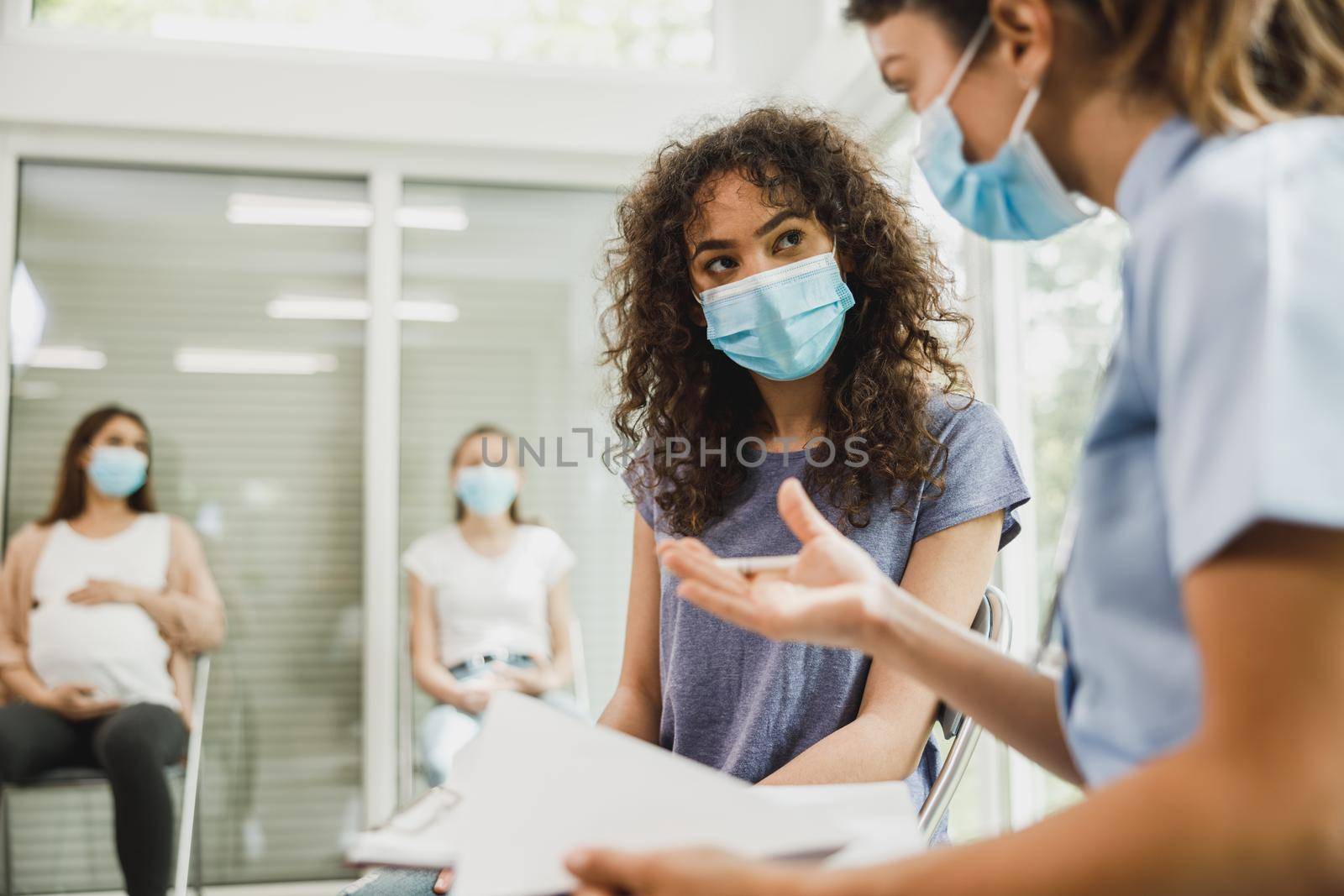 Upset African American teenage girl with protective mask talks with nurse while waits for gynecologist check up.