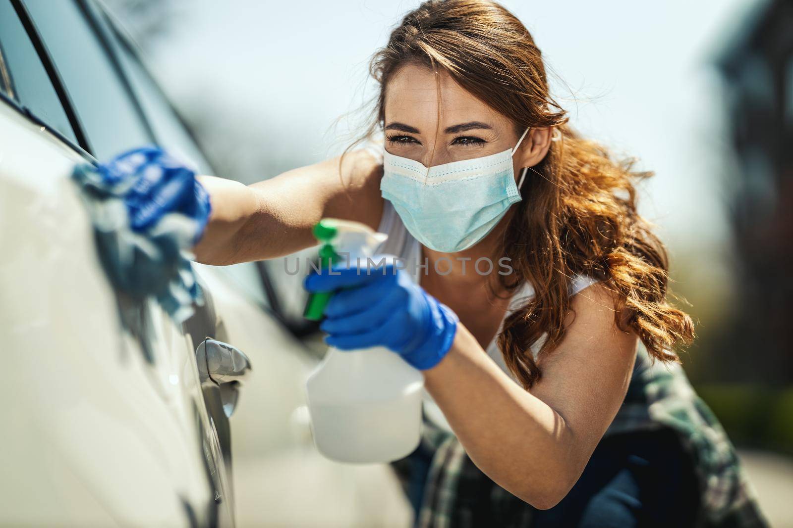 A young woman with a mask on her face and protective gloves on her hands, wipes her car holding a cloth in one and a bottle with disinfectant in the other hand.