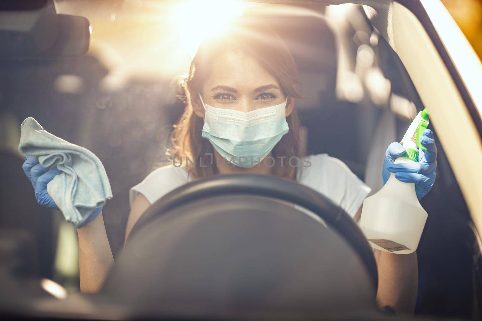 A young woman with a mask on her face  and protective gloves on her nands is ready to wipe her car holding a cloth in one and a bottle with disinfectant in the other hand.
