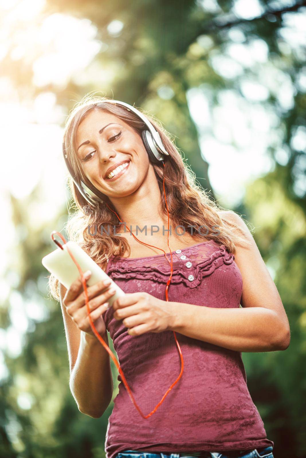 Woman relaxes with headphones listening to music sitting in park. She is doing selfie on her smartphone