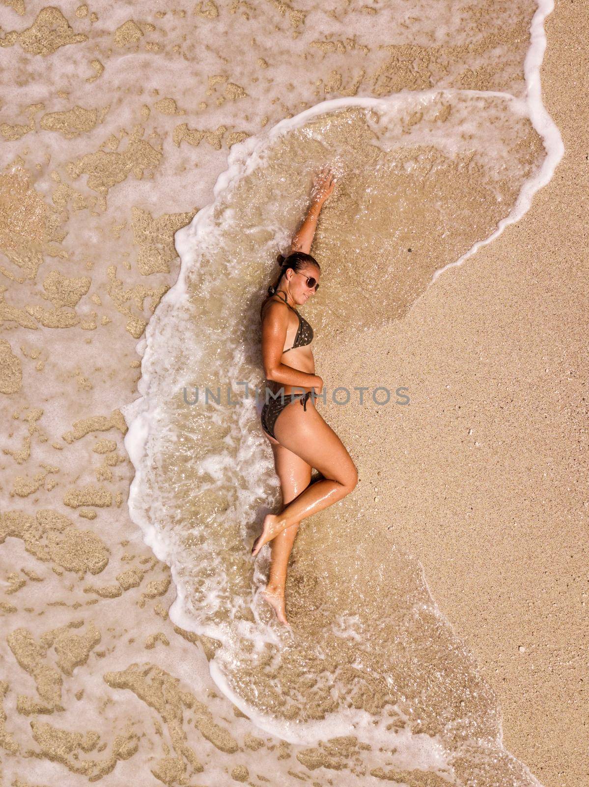 Aerial view picture from drone of woman relaxing on the idyllic tropical beach. She is laying on the sandy beach while the waves splash on it. 