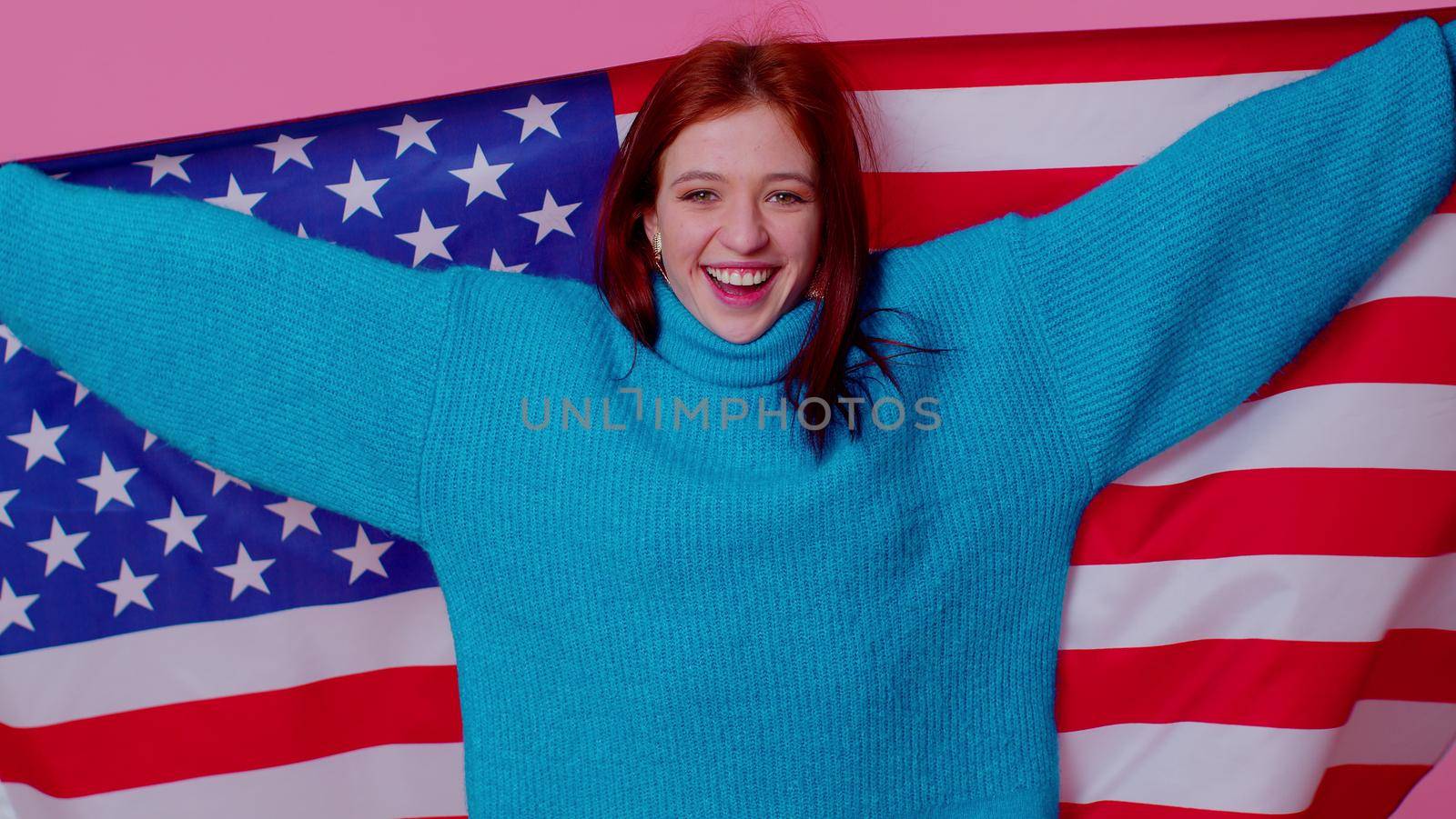 Trendy cheerful positive teen girl waving and wrapping in American USA flag, celebrating, human rights and freedoms. Independence day. Young pretty adult woman. Indoor studio shot on pink background