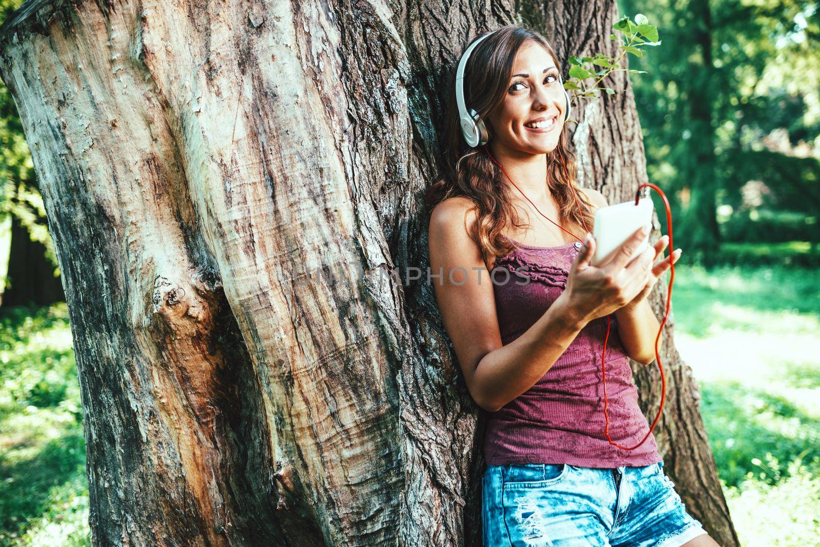 Smiling beautiful modern woman listening to music with phone standing under a tree in a city park 