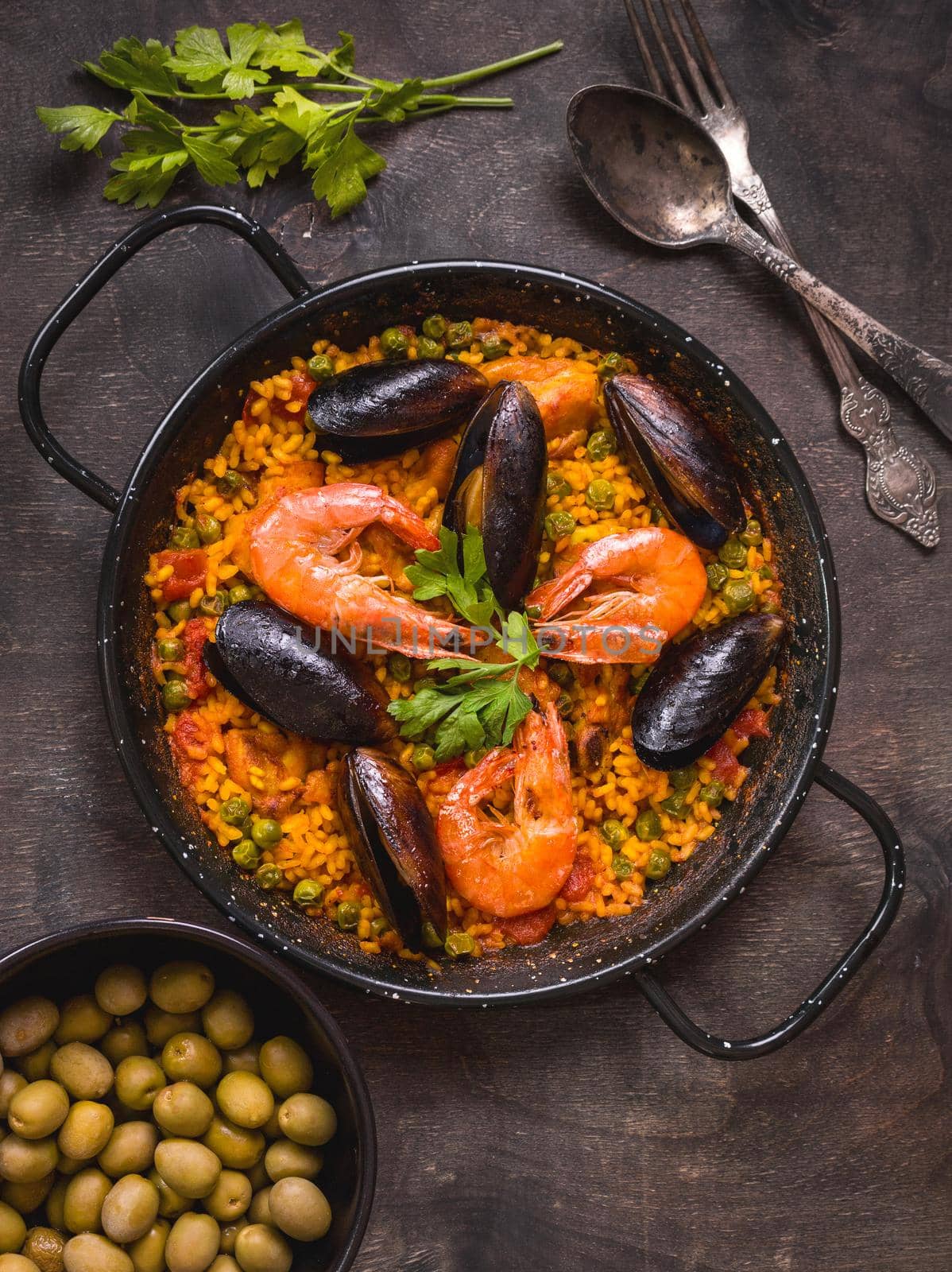 Paella in black pan with rice, shrimps, mussels, squid and meat, bowl with olives and vintage cutlery. Seafood paella, traditional spanish dish. Paella on rustic table. Selective focus. Top view