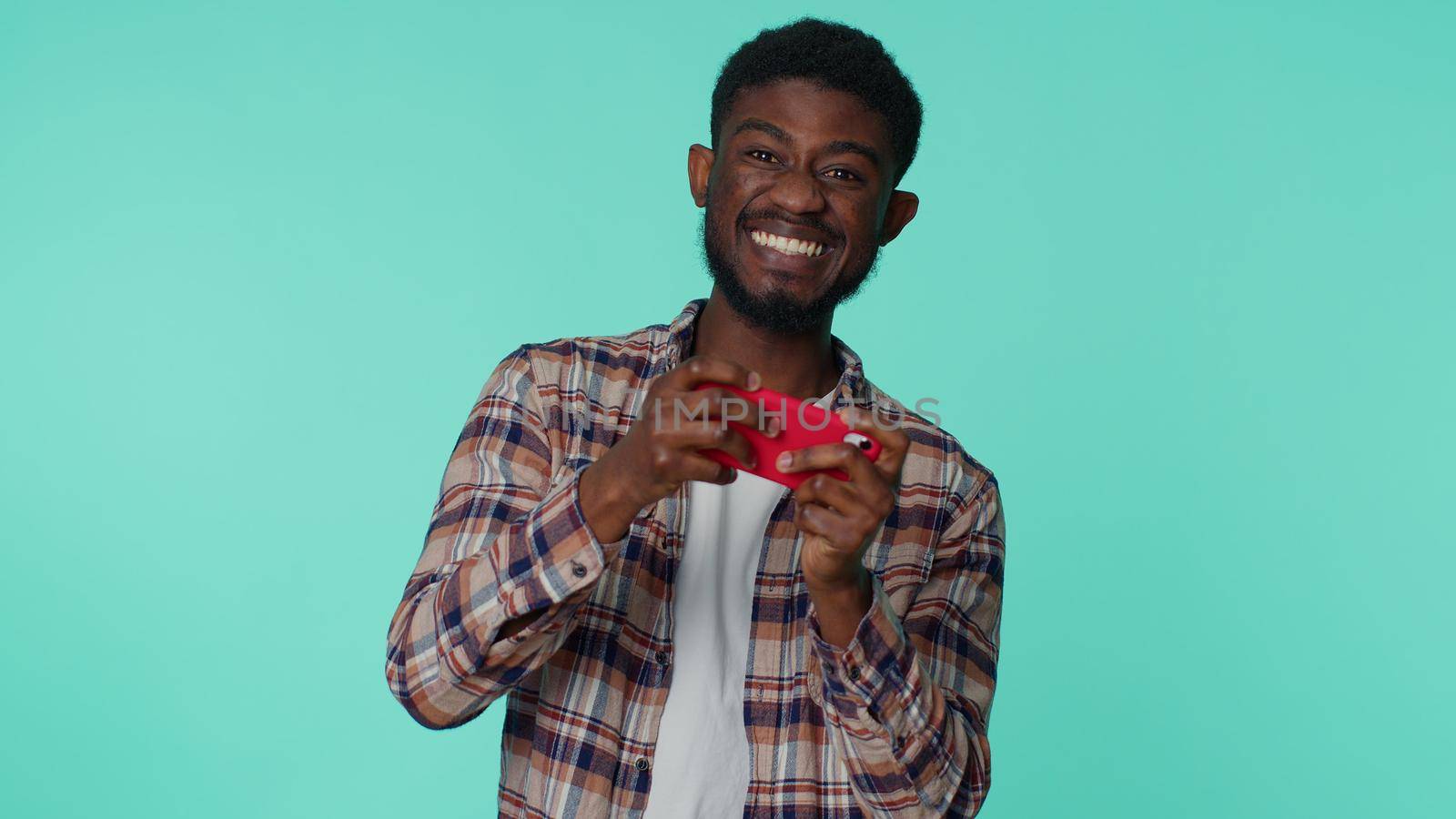 Worried funny addicted african american man enthusiastically playing racing video games on mobile phone. Young guy using smartphone gadget app with drive simulator isolayed on blue studio background