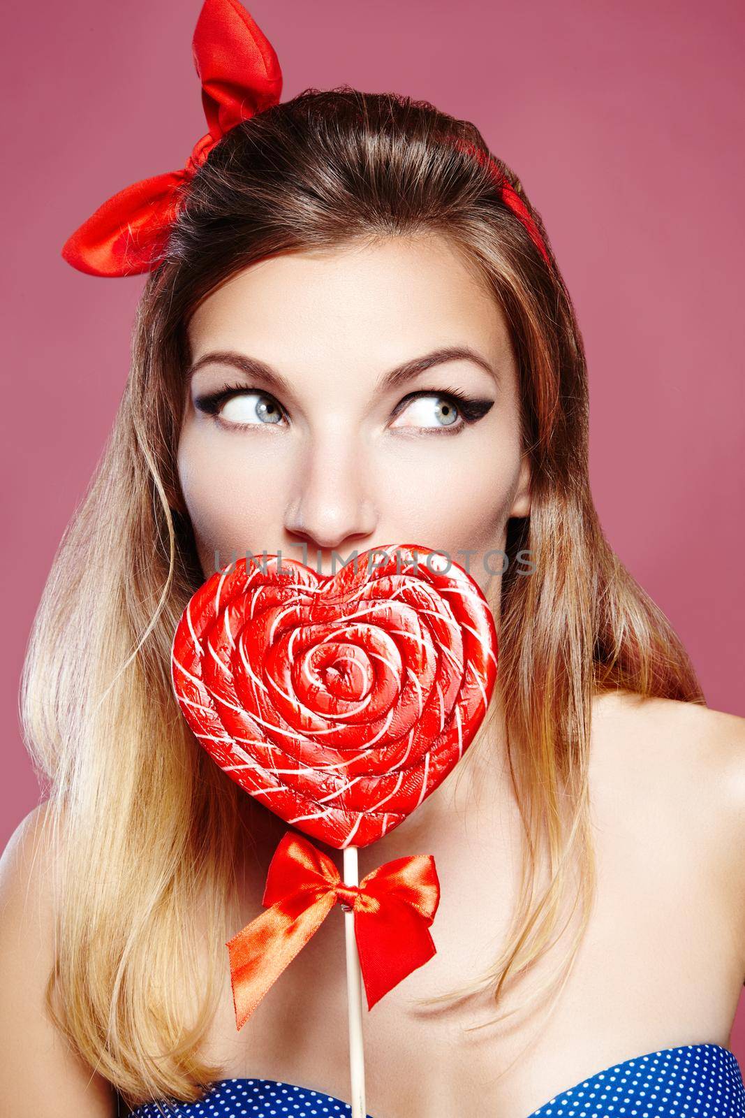 Sexy american style. Pinup girl with cute red bow accessories in her blond hair. Playful happy young woman with sweet candy lollipop in heart symbol shape. Beautiful valentine style of beauty