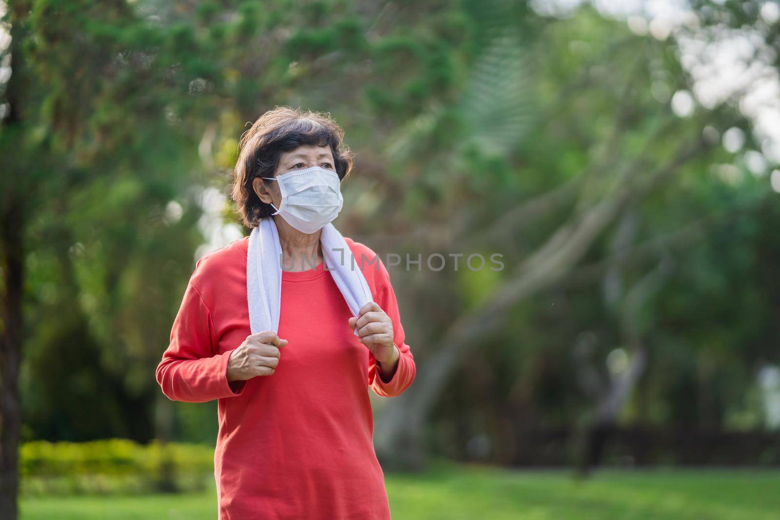 Athletic asian senior woman 60s wearing surgical mask and jogging. Beautiful senior asian woman running at the park on a sunny day.