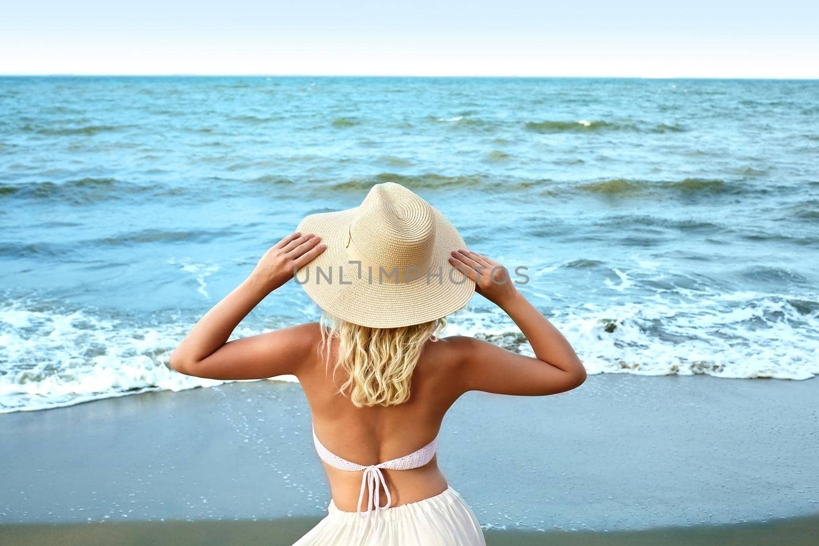 Blonde Woman in Fashion Summer Style Standing at Sea and Holding Hat. Luxury Lifestyle Rear View. Summer Vacation Concept with Ocean. Soul Relaxing, Spirit Calm