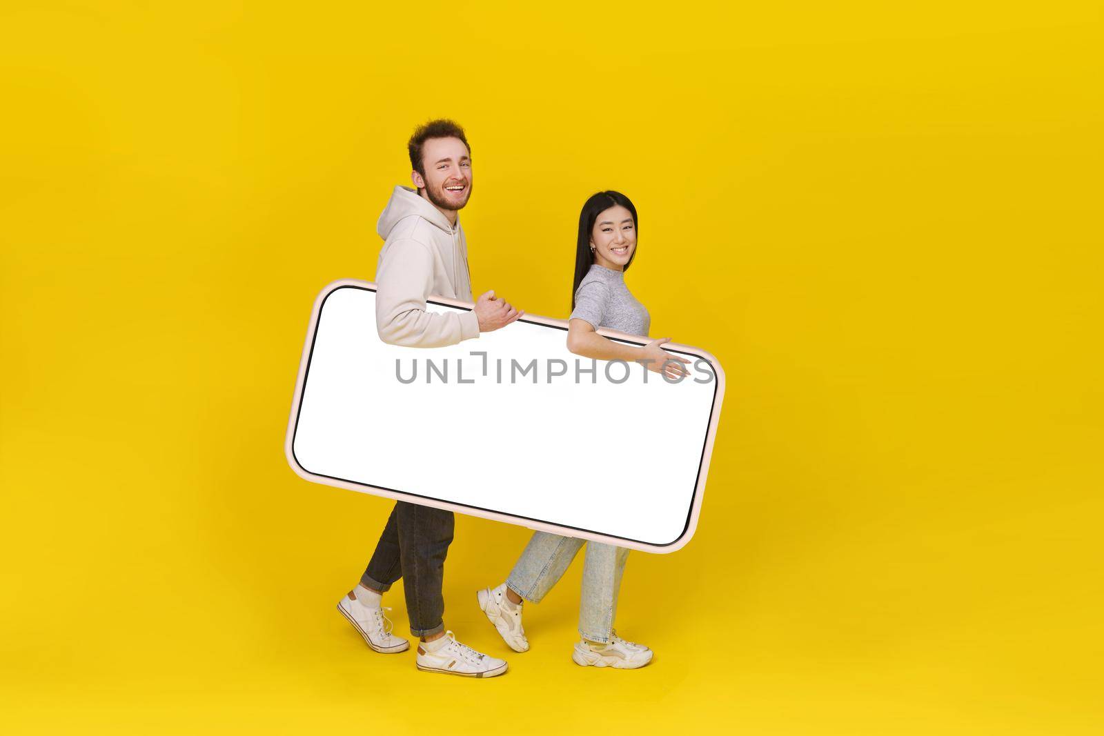 Asian girl and caucasian guy holding huge smartphone showing blank white screen, mobile app advertisement and excited smile on camera isolated on yellow background. Product placement.