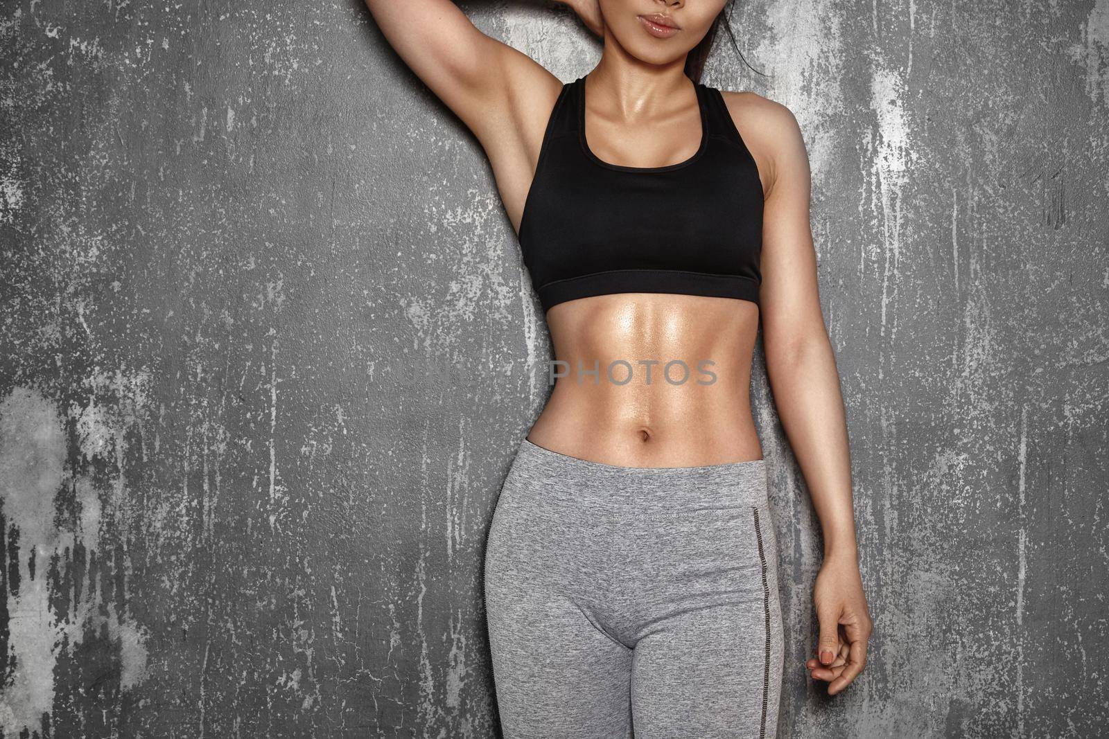 Fit Woman style with sexy hips. Female with perfect abdomen muscles. Beautiful Body in sport clothes near grey wall.