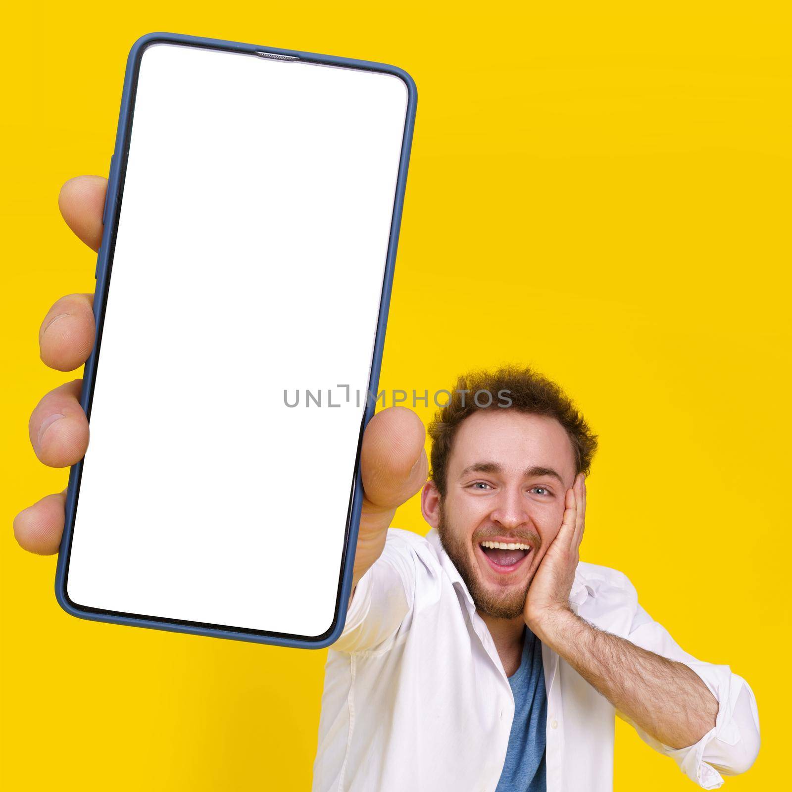 Great offer. Young happy man holding smartphone showing a white empty screen and exciting to win isolated over yellow background, celebrating success. Product placement for mobile app advertisement.