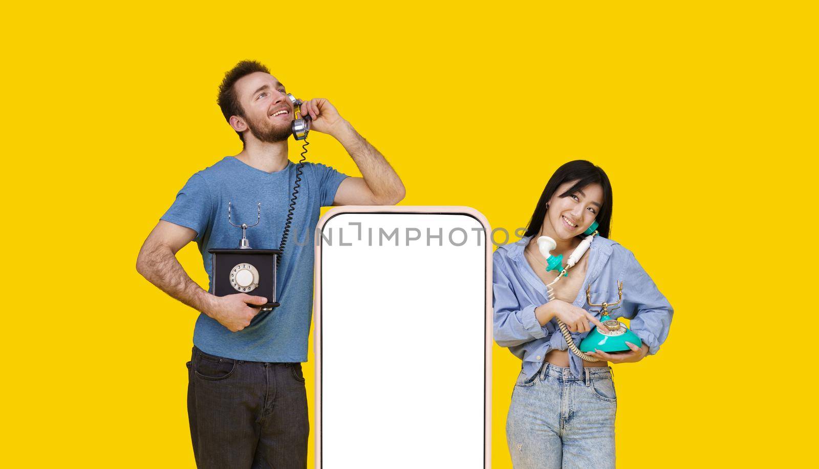 Asian girl and caucasian guy in love talking on vintage phones leaned on huge smartphone with blank screen, looking at each other isolated on yellow background. Product placement by LipikStockMedia