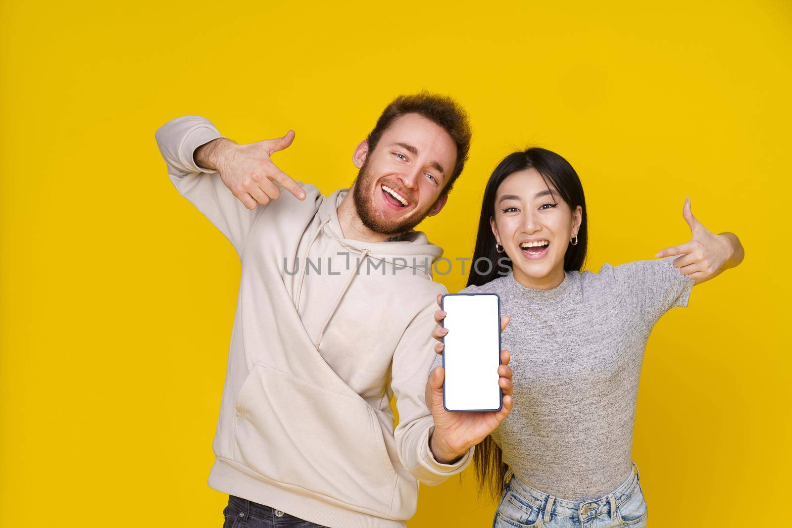 Asian girl and caucasian guy showing thumbs up holding smartphone showing blank white screen, mobile app advertisement and excited smile on camera isolated on yellow background. Product placement.
