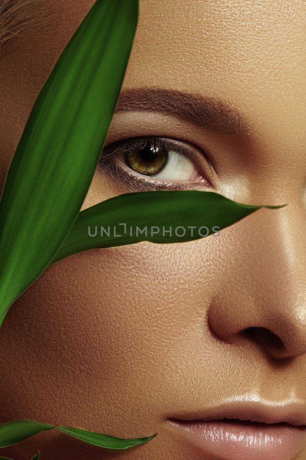 Close-up face of beautiful young woman with smooth tanned skin covered by leaves. Portrait of beauty model with natural bronze make-up. Spa, skincare and wellness